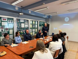 na dużej sali przy stole siedzą kobiety wśród nich siedzą umundurowani policjanci jeden policjant stoi widać również duży ekran