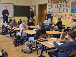 klasa szkolna , siedzące dzieci przy ławkach dwóch umundurowanych policjantów stoi  na środku klasy za ich plecami widoczna jest duża tablica