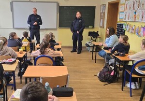 klasa szkolna , siedzące dzieci przy ławkach dwóch umundurowanych policjantów stoi  na środku klasy za ich plecami widoczna jest duża tablica