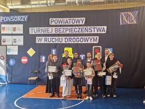 na sali stoją dzieci trzymając dyplomy w rękach za ich plecaki stoi policjantka oraz dwaj mężczyźni i kobieta
