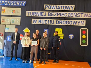 na sali stoją dzieci trzymając dyplomy w rękach za ich plecaki stoi policjantka oraz dwaj mężczyźni i kobieta