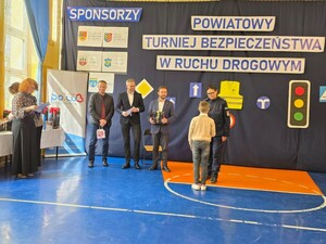na sali stoją dzieci trzymając dyplomy w rękach za ich plecaki stoi policjantka oraz dwaj mężczyźni i kobieta