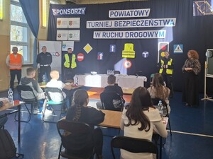 policjanci mężczyzna w odblaskowej kamizelce kobieta trzymająca kartkę w rękach za plecami stojących osób duży napis  turniej bezpieczeństwa w ruchu drogowym , siedzące dzieci przy ławkach