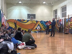 na sali gimnastycznej siedzą na podłodze harcerze przed nimi stoją dwaj policjanci za nimi koło szklanych drzwi stoją dwaj harcerze i jedna harcerka