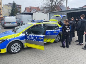 młodzież stoi wokół radiowozu który ma pootwierane drzwi i bagażnik