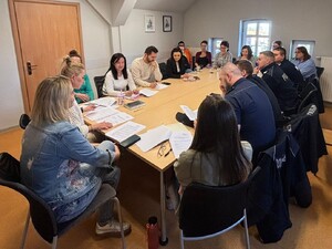 widocznie są osoby siedzące w  sali przy dużym stole z  jednej strony siedzą umundurowani policjanci z drugiej pracownicy miejskiego ośrodka pomocy społecznej