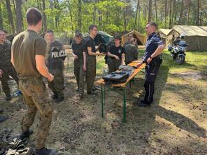 las, polana namiot przy ławce stoi policjant na ławce leżą sprzęty policyjne widoczny jest motocykl