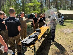 las, polana namiot przy ławce stoi policjant trzyma tarczę w ręku na ławce leżą sprzęty policyjne widoczny jest motocykl
