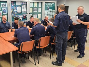 policjanci siedzą na krzesłach wokół stołu obok stoją policjanci trzymający w ręku kartki papieru