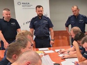 przed stołem stoi trzech policjantów widoczni są również siedzący policjanci przy stole