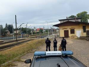 trzech umundurowanych funkcjonariuszy oraz pies  jeden policjant i dwóch z Straży Ochrony Kolei w tle tory i dworzec oraz samochód służbowy